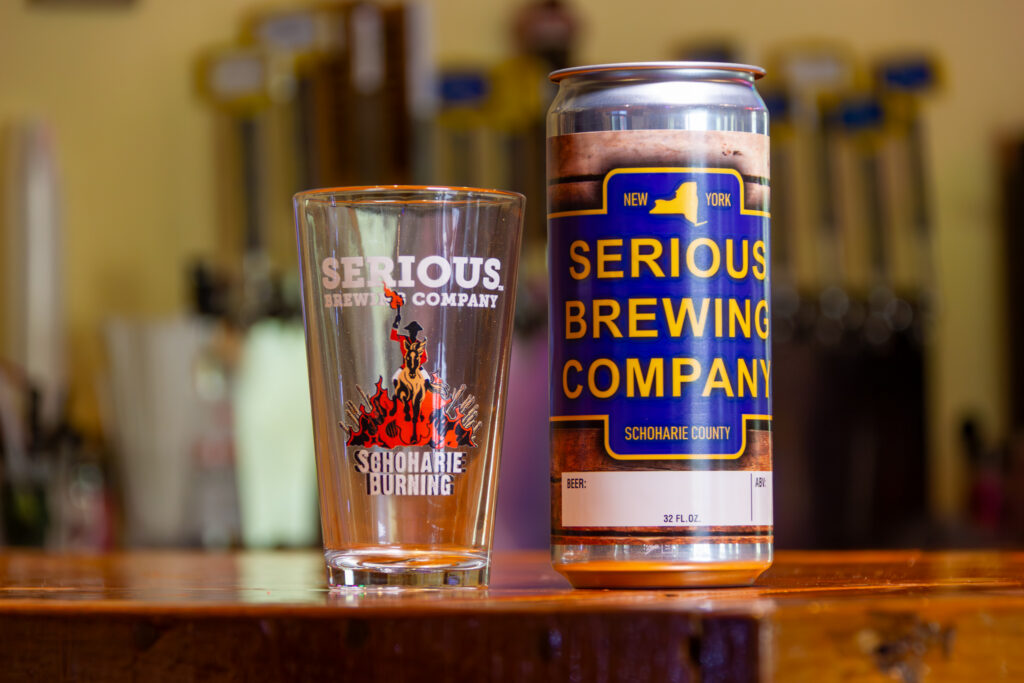 "Schoharie Burning" beer in a can, next to a glass with artwork of a man riding on a horse with flames in the background.