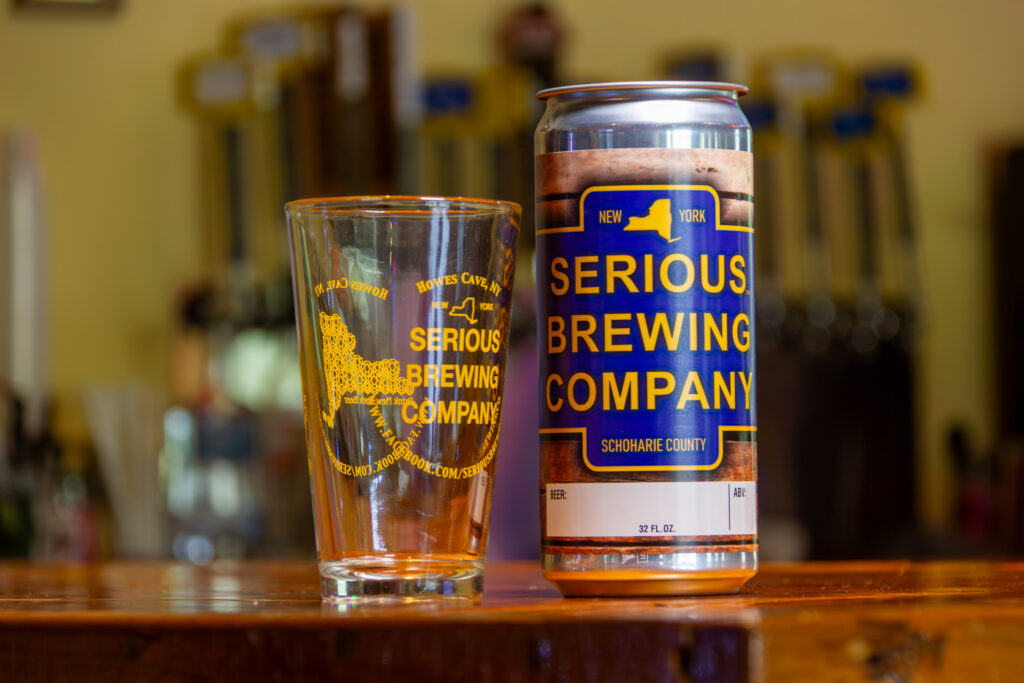 "Serious Blonde" beer in a can, next to a glass with the Serious Brewing logo.