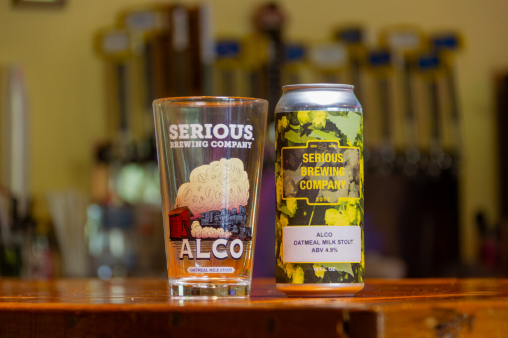"Alco Oatmeal Milk Stout" beer in a can, next to a glass with artwork of a train blowing smoke.